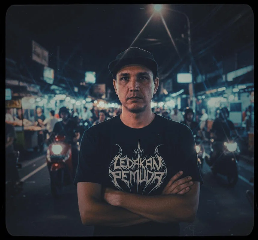 Man in a flat cap with arms crossed, wearing a Ledakan Pemuda shirt on a night street in Jakarta with motorbikes behind him